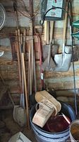 Wall-mounted metal gardening tools including multiple shovels and rakes alongside wooden handles visible