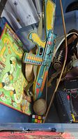 Photo showing a colorful toy rifle with a strap, Hardy Boys game board partially visible, toy bow, and other items stacked in a bin