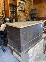 Side angle of the black wooden chest inside a workshop or storage building, showing the base and side of the chest.