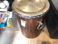 Overall view of the wood bin with metal lid and ring handles placed on a floor.
