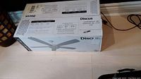 Photo showing the Monte Carlo Discus ceiling fan original box, lying on floor, labeling visible with model and dimensions.