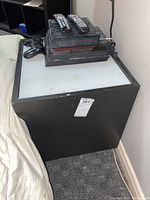 Black manufactured wood cube with a white, glass-like top showing scratches and wear. Items including remote controls and a gaming controller on top, not included.