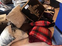 Group of nine cushions displayed on a table, showing various sizes and patterns including plaid, fuzzy, animal motifs, and plain designs.