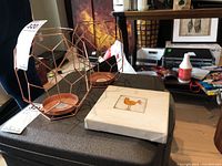 Two metal geometric candle holders with a copper tone, small framed bird print on a white mat, and part of a copper metal wall sconce.