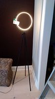 Full view of the floor lamp, showing the illuminated circular ring light and the black metal tripod base against a brown and white wall background.