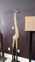 Full view of the tall wooden giraffe sculpture against a dark wall, showing the overall appearance and painted pattern.