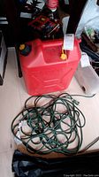 Red plastic Jerry can with black cap and yellow vent, next to coiled green extension cord on floor.