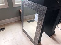 Mirror with decorative metal mosaic frame positioned on floor near window, showing full front angle with natural lighting.
