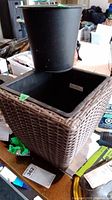 Photo showing the round black resin pot placed on top of the square woven resin planter with black liner inside, on a cluttered indoor surface.