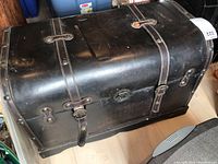 Black vinyl vintage trunk with two leather buckle straps across the top and metal rivets, shows wear and stains on the surface.