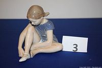 Front-side view of the porcelain figurine showing the girl sitting with one leg bent putting on a ballet shoe, wearing a blue dress.