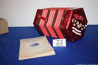 Concertina with booklet in front, showing red finish, buttons, straps, and booklet cover