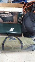 Coleman propane stove green metal case, Ozark Trail tent carrying bag, rolled sleeping bag bag in blue and green fabric.