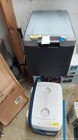 View of two coolers: one blue and white standard cooler with cup holders on lid, one dark gray plug-in travel cooler with textured lid.