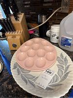 Photo of a pink ceramic egg dish placed on a round ceramic platter with a blue-gray leaf motif, with a partial view of the wooden knife block nearby.
