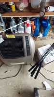 Beige Lasko heater positioned on floor next to shelf with fluids and miscellaneous items.