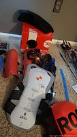 Photo of orange Rossignol ski race helmet on its branded box with ski goggles, shin guards, and white-gray chest protector visible on carpet.