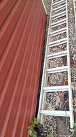 Aluminum extension ladder laid out on the ground along a red metal building exterior, showing length and condition.