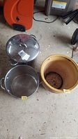 Wide shot showing the ceramic planter and two cooking pots, one with lid, all on concrete floor with some dirt and debris