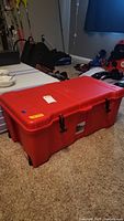 Red plastic Contico Ultra Storage Bin closed on carpeted floor with visible black latches and handles, surrounded by sports equipment.