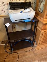 Photo of the wood-toned three-shelf printer stand with a white printer and black keyboard on top, cords visible underneath, showing the general condition and design.