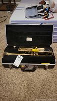Open view of brass trumpet in black lined hard case on carpeted floor, tag Number 211 attached to trumpet.