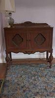 Front view of wooden accent cabinet with two decorative doors and carved details on the bottom apron. Shows scratches and wear on surface.