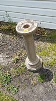 Side view of tall concrete bird bath stand with peeling top surface and hexagonal base in outdoor ground setting.