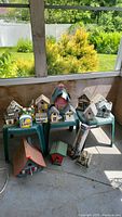 Photo showing a collection of approximately 15 decorative bird houses arranged on green plastic stools, varying in style, color, and size from small to large, alongside one ceramic napkin holder.