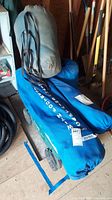 Three sleeping bags in carry bags, including grey and blue bags, stacked in a corner near garden tools.