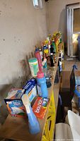 Photo of assorted cleaning supplies including liquid detergents, fabric softeners, spray cleaners, and boxed dryer sheets arranged on a wooden shelf against a concrete wall.