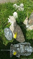 Concrete duck, resin angel figurine, squirrel figurine, and round shell-shaped garden piece displayed on grass with visible dirt and weathering.