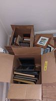 Two cardboard boxes filled with assorted picture frames of different sizes and materials, including wooden, metal and plastic frames. The frames vary in color and finish, mostly in 4x6 and 8x10 sizes.