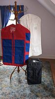Photo shows 22-inch black Samsonite wheeled carry-on suitcase placed under wooden coat rack, supporting three garment bags: two red and blue Wrap Pack garment bags and one white Saks Fifth Avenue garment bag.