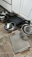 Wide view of pots, pans, grill plate, mixing bowls stacked, and perforated baking sheet arranged on kitchen floor.
