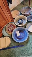 The photo shows a group of pottery bowls and platters arranged on carpet. Includes bowls of various sizes and colors, a covered pot, a glass bowl, and a large Pfaltzgraf blue glazed bowl.