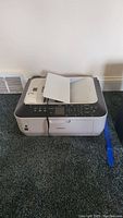 Canon Pixma MX330 all-in-one printer on carpet floor, closed scanning lid, paper feeder open with paper inside.