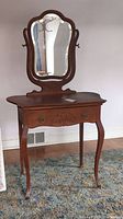 Front view of wooden vanity showing single drawer with brass handles, scalloped front edge, and curvy cabriole legs. Adjustable, bevelled edge mirror mounted on back.