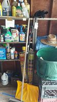 Photo shows shelf with assorted garden chemicals, sprays, paint supplies, broom, rolled weed block fabric, garden shovel, and straw hat beside a collapsible garden basket.