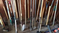 Wide view showing a collection of yard tools arranged against a wooden wall featuring shovels, forks, pickaxes, and sledgehammer.