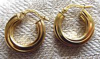 Close-up photo showing both earrings from the front on textured fabric, highlighting the thick rounded hoop shape with a polished yellow gold finish and visible clasp mechanism.