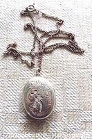 Oval sterling silver locket showing intricate floral engraving on front and attached chain.