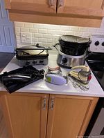 Photo shows Hamilton Beach 3-bowl crockpot with ceramic inserts, Waring electric handheld mixer, Robo Twist electric jar opener, Cuisinart Griddler and a black Hamilton Beach electric appliance on a kitchen counter.