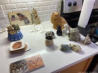 Photo of the entire collection including various cat statues, coasters, and figurines on a kitchen counter with tile backsplash