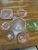 Eight colored glass serving pieces arranged on a wood table, showing pink and green glass bowls and one serving platter with decorative embossed patterns.