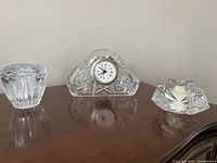 Three crystal items on wooden surface: one vintage crystal mantel alarm clock and two votive candle holders.