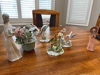 Six porcelain figurines arranged on a table: lady with kitten in white dress, mint green pot with glass flowers, two bird figurines on floral base, water bird, lady in pink with bouquet.