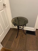 Full view of round smoked glass top accent table positioned against wall and door, showing metal base with tripod legs and rope detail.