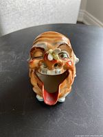 Front view of ceramic ashtray shaped as man's face, with tongue-like mouth opening and bee on nose.