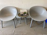 Two grey plastic outdoor tub chairs with matching grey side table with boxed teacup planter on top, against a beige wall.